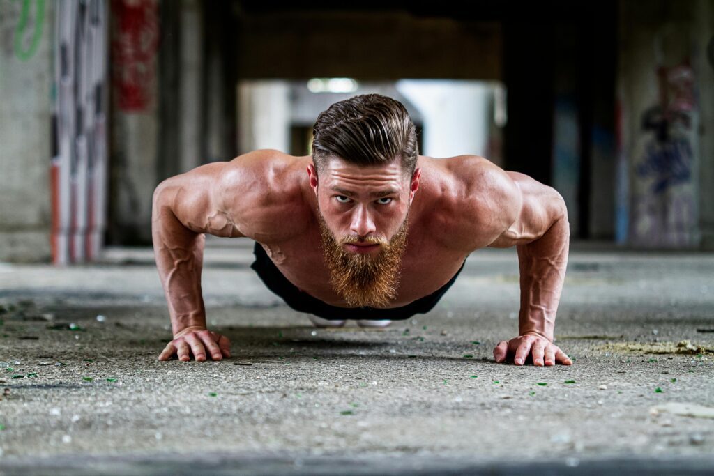 Fit shirtless man with a beard doing a push-up exercise on concrete floor.